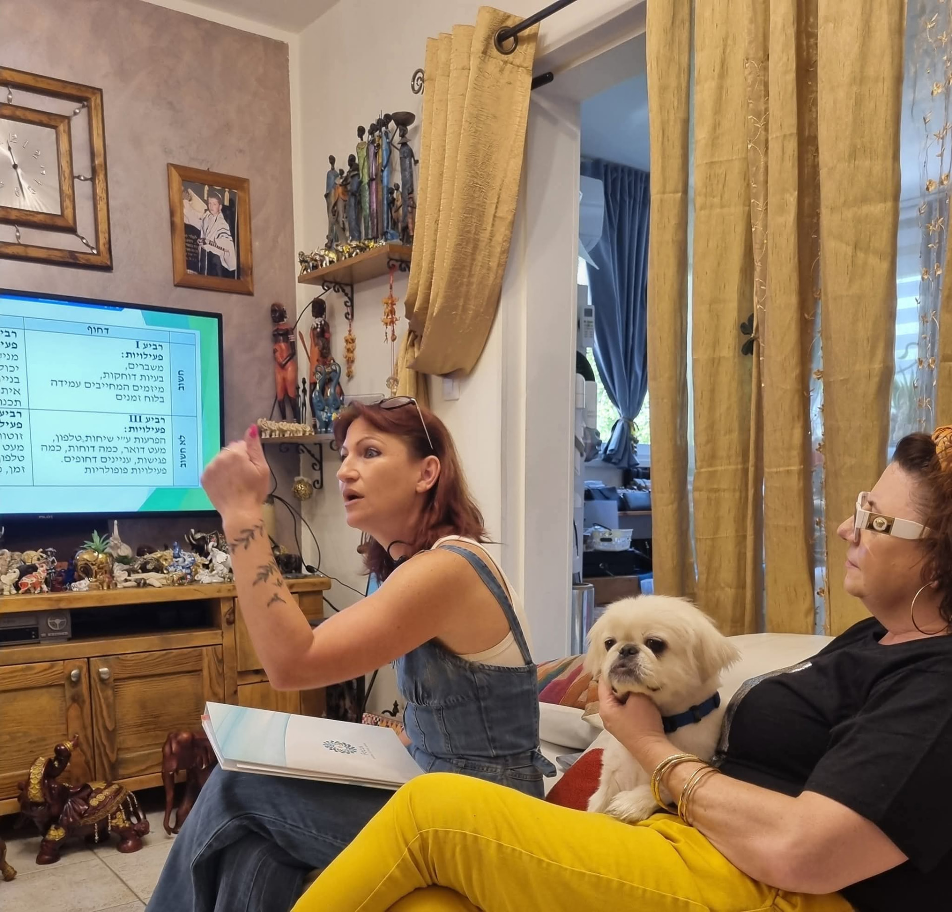 לודמילה מנחה שיחת ליווי בסלון ביתי, מול אישה השומעת על הספה ועם כלב לבן קטן על ברכיה
