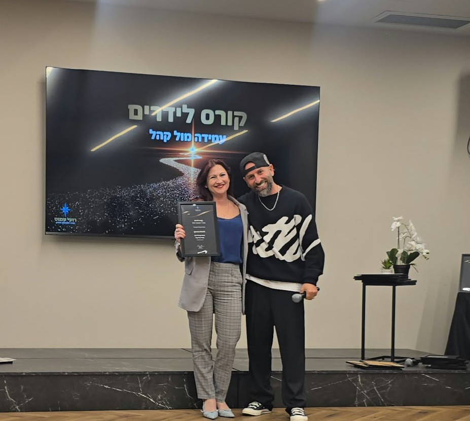 לודמילה עומדת על במה ומחזיקה תעודת סיום בקורס עמידה מול קהל, לצדה מנחה המחזיק מיקרופון